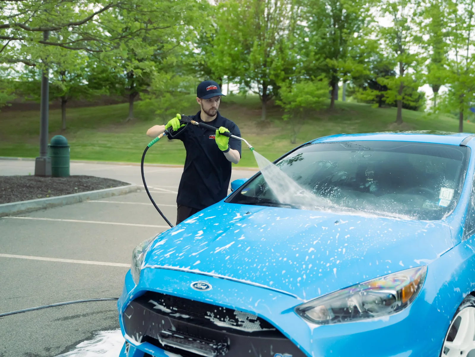 Detailer in a black uniform pressure-rinsing foam off the hood and windshield of a blue Ford Focus RS in a parking lot