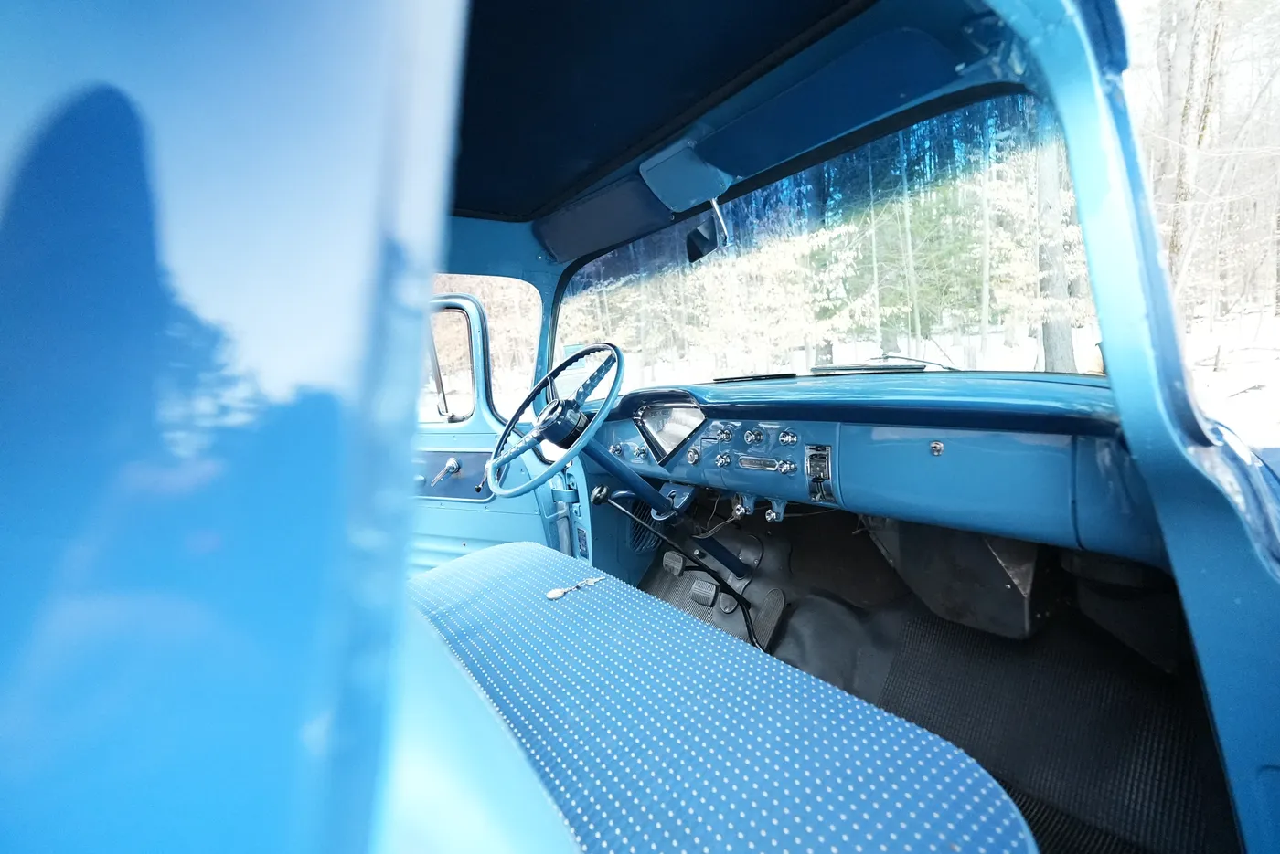 Classic pickup cab seen past the open blue passenger door, showing the dotted bench seat, dash gauges and steering wheel