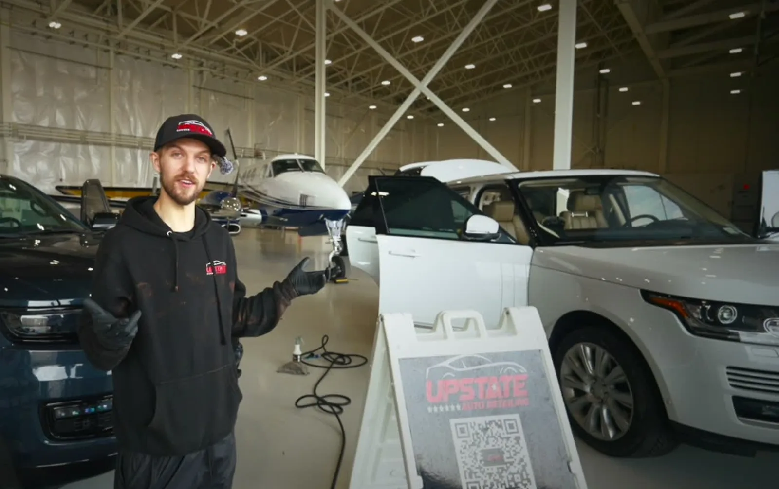 Detailer in an Upstate cap beside the Upstate A-frame sign, two SUVs and a twin-prop plane parked in a hangar