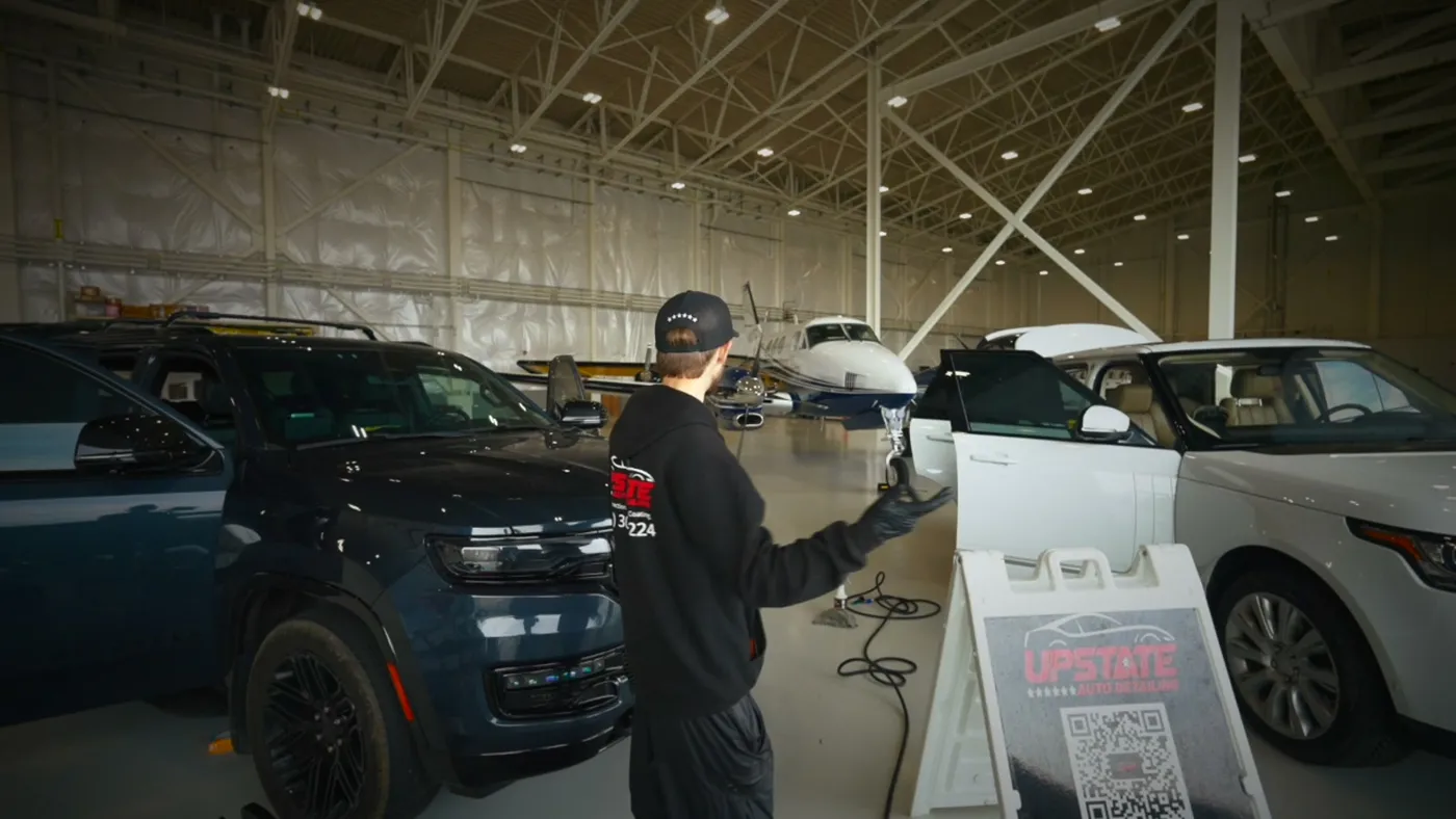 Detailer seen from behind gesturing at a white SUV with its door open, small plane parked further back in the hangar