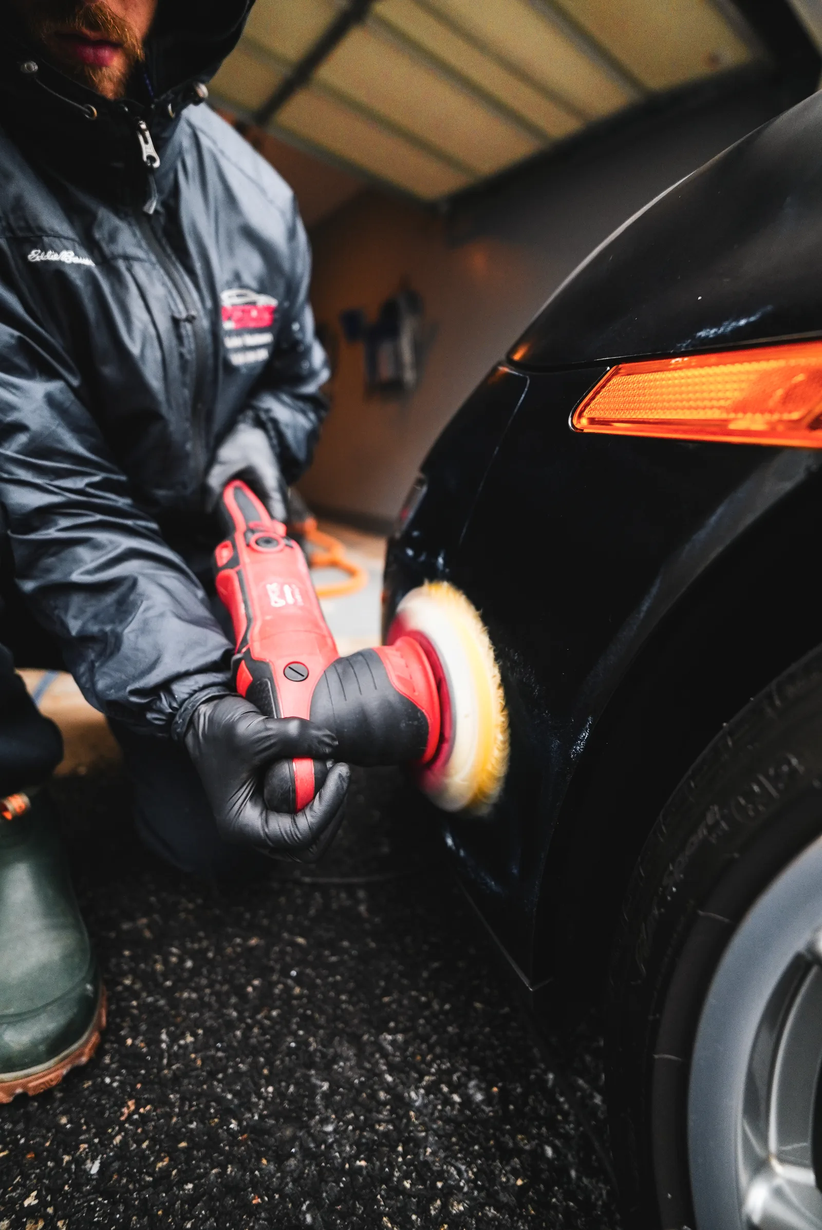 Detailer polishing the front fender of a black car with a red machine polisher and wool pad in a garage
