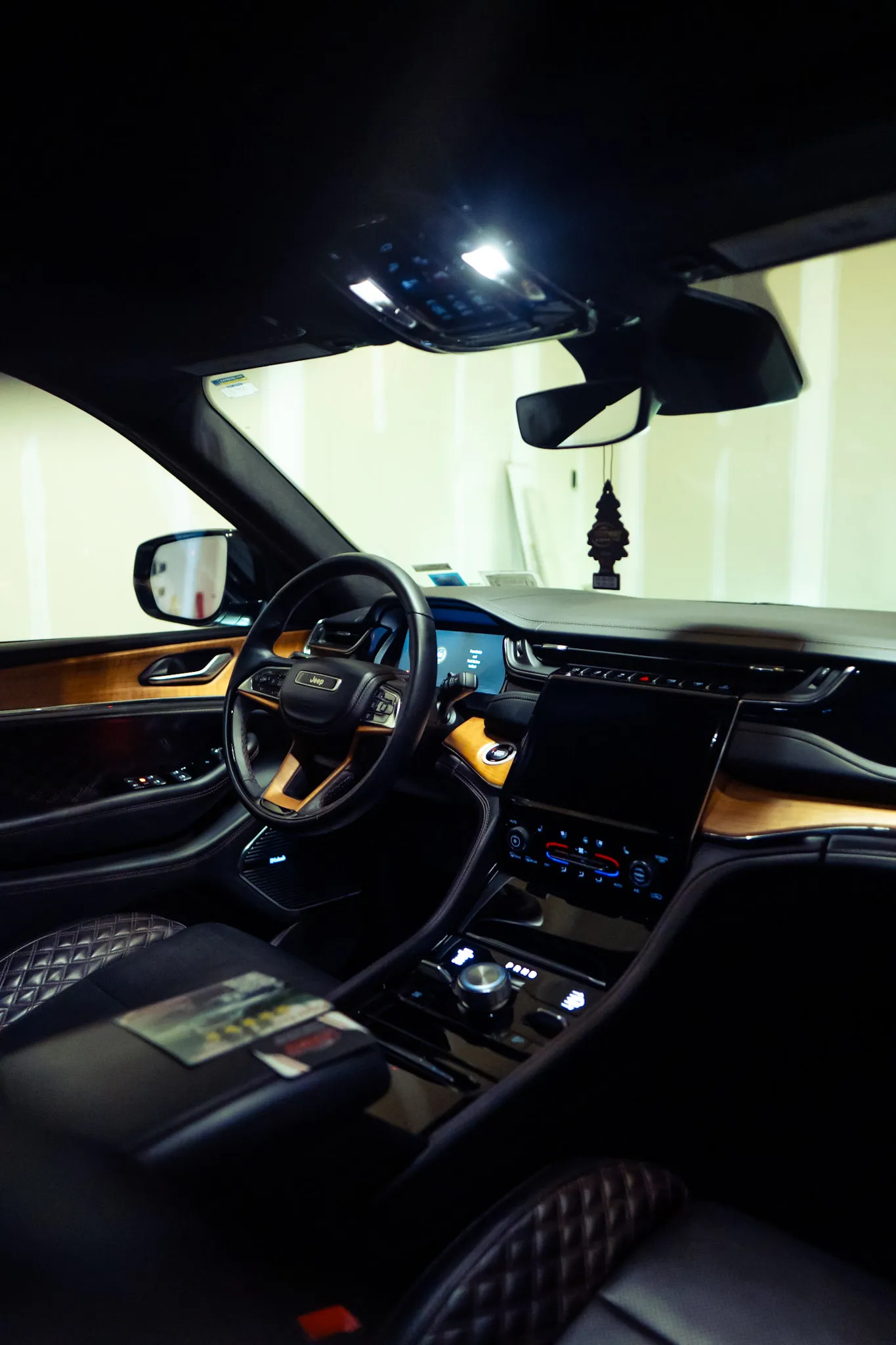The dashboard, center screen and wood trim of a Jeep interior lit by the dome light