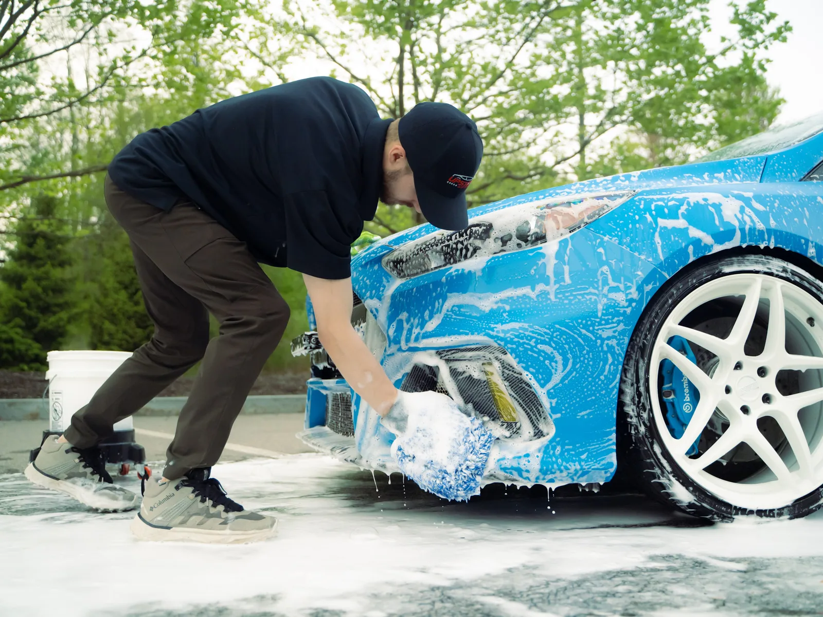 A detailer hand washing the soapy front bumper of a blue Ford Focus RS with a wash mitt, a bucket behind him