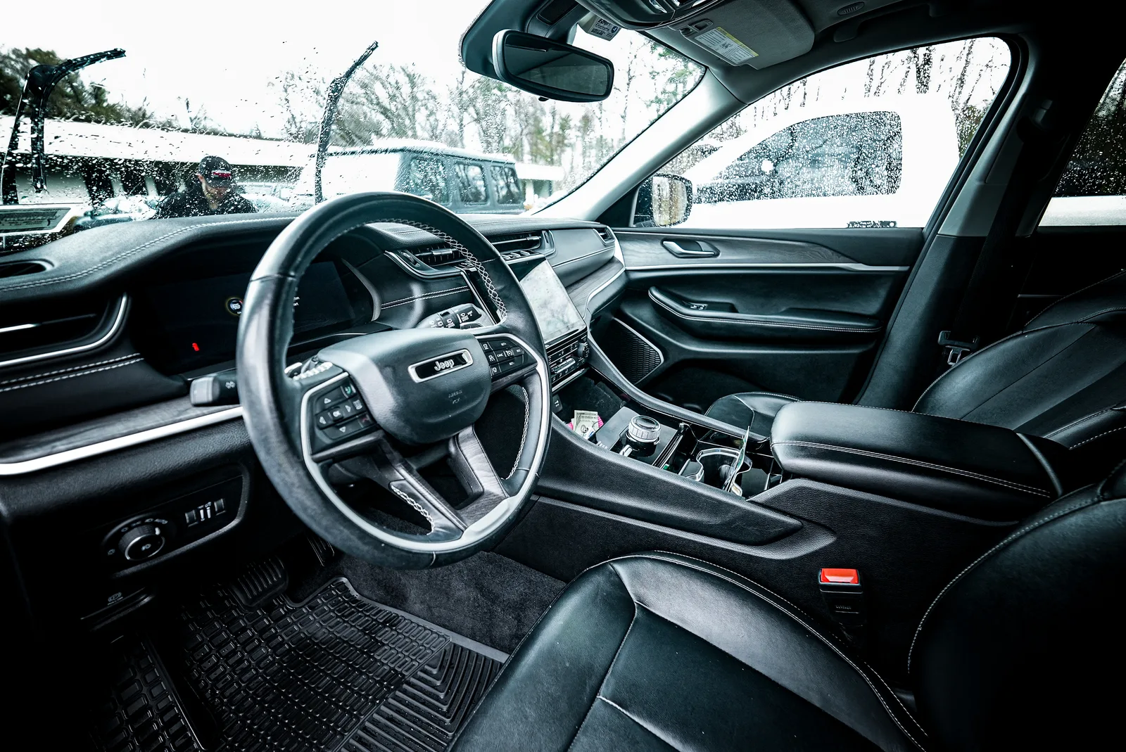 Front seats, steering wheel and dash of a Jeep with black leather seats and rubber floor mats, a detailer visible through the windshield