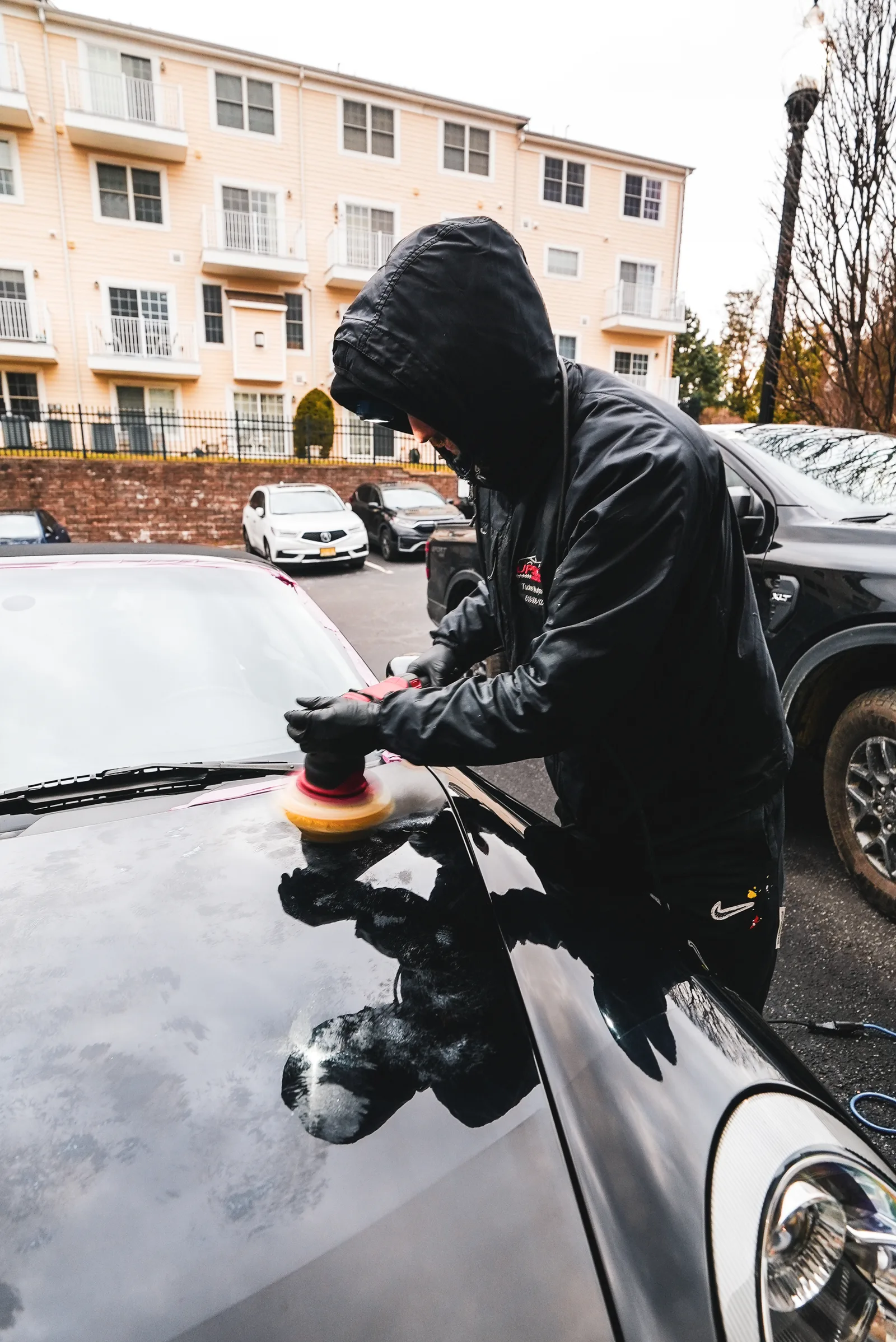 A detailer in a black hooded jacket machine polishing the hood of a black car in a parking lot